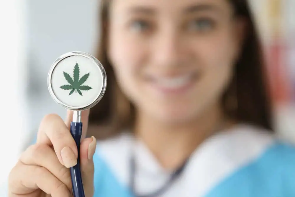 CBD un toccasana per la pressione alta. Una figura femminile sorridente sullo sfondo esibisce in primo piano un fonendoscopio sul cui disco si trova una piccola foglia di canapa.