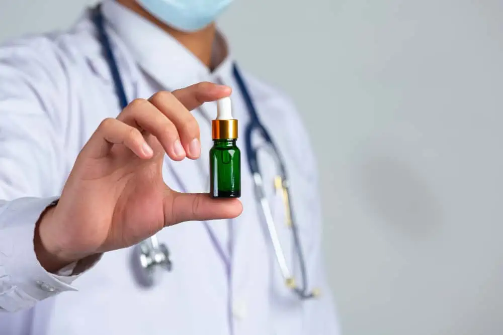La figura sfumata di un sanitario con camice bianco, mascherina e fonendoscopio al collo, mostra in primo piano la mano destra con un piccolo flacone in vetro verde con un contagocce dorato.