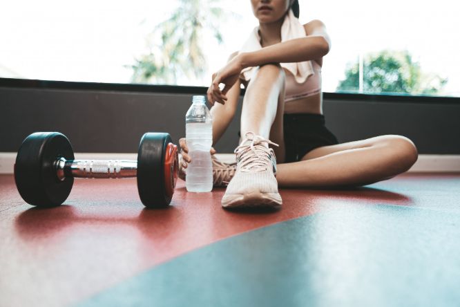 Olio CBD per crampi e dolori post workout. L'immagine mostra una giovane donna in abbigliamento da palestra mentre si accinge a bere da una bottiglia di plastica. Sul pavimento a riquadri rossi e blu, un manubrio nero e rosso.